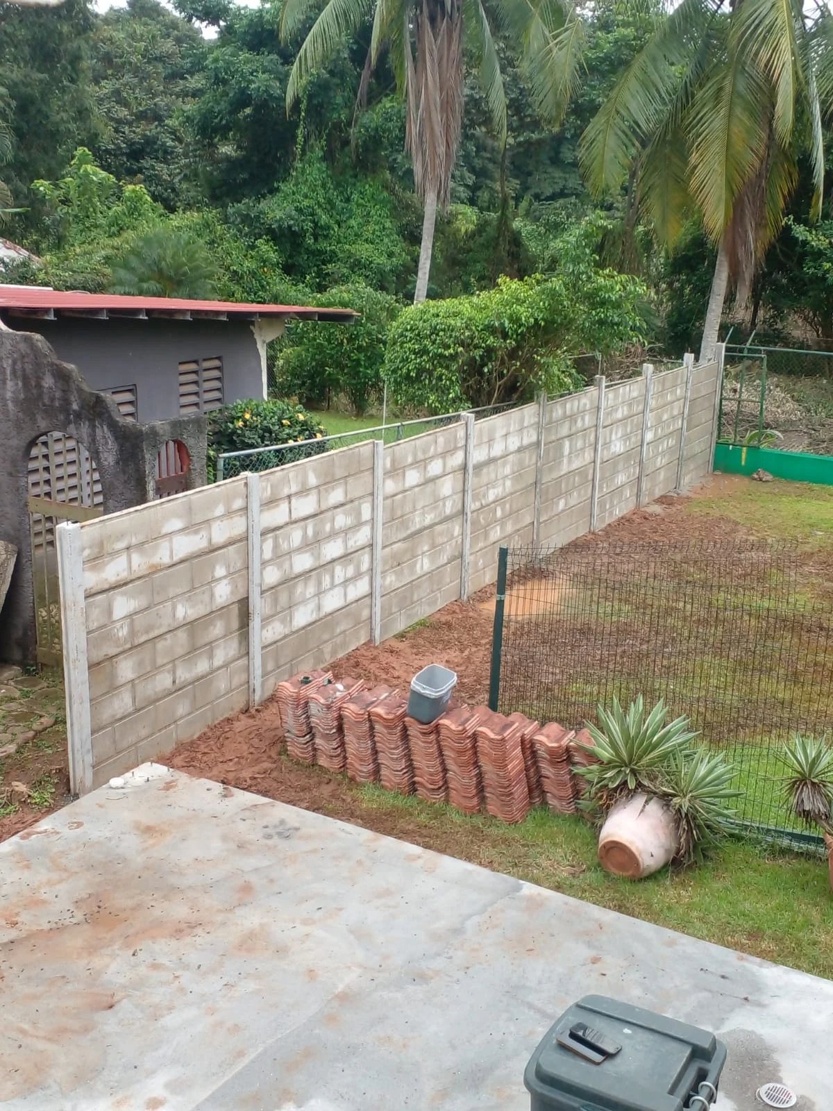 Muro perimetral prefabricado terminado dividiendo lote junto a tejas tradicionales apiladas