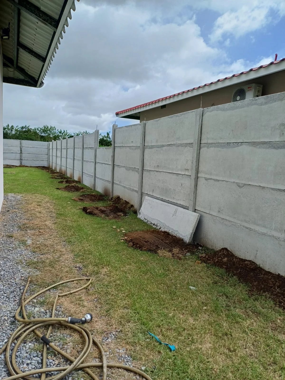 Muro perimetral prefabricado lateral con manguera en el jardín y vivienda con aire acondicionado