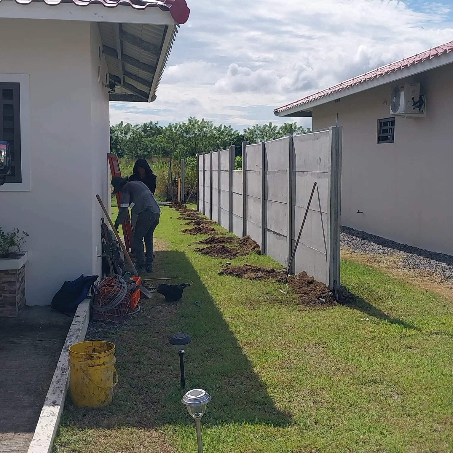 Cuadrilla instalando muro perimetral lateral junto a vivienda con techo de teja y poste solar
