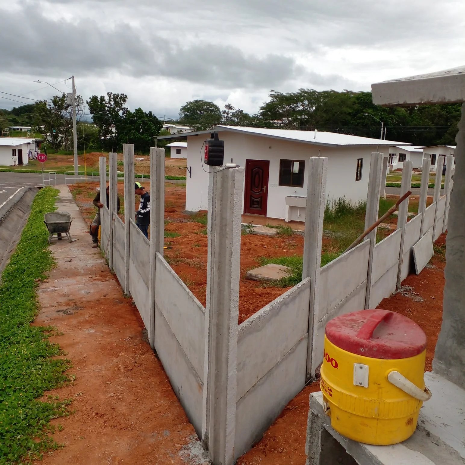 Muro perimetral prefabricado en U delimitando lote con vivienda y poste de luz al fondo