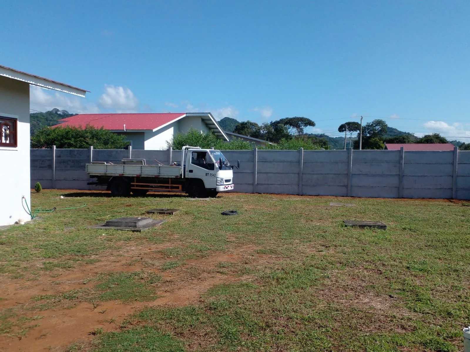 Vista lateral de muro perimetral prefabricado con camión y vivienda con techo rojo al fondo
