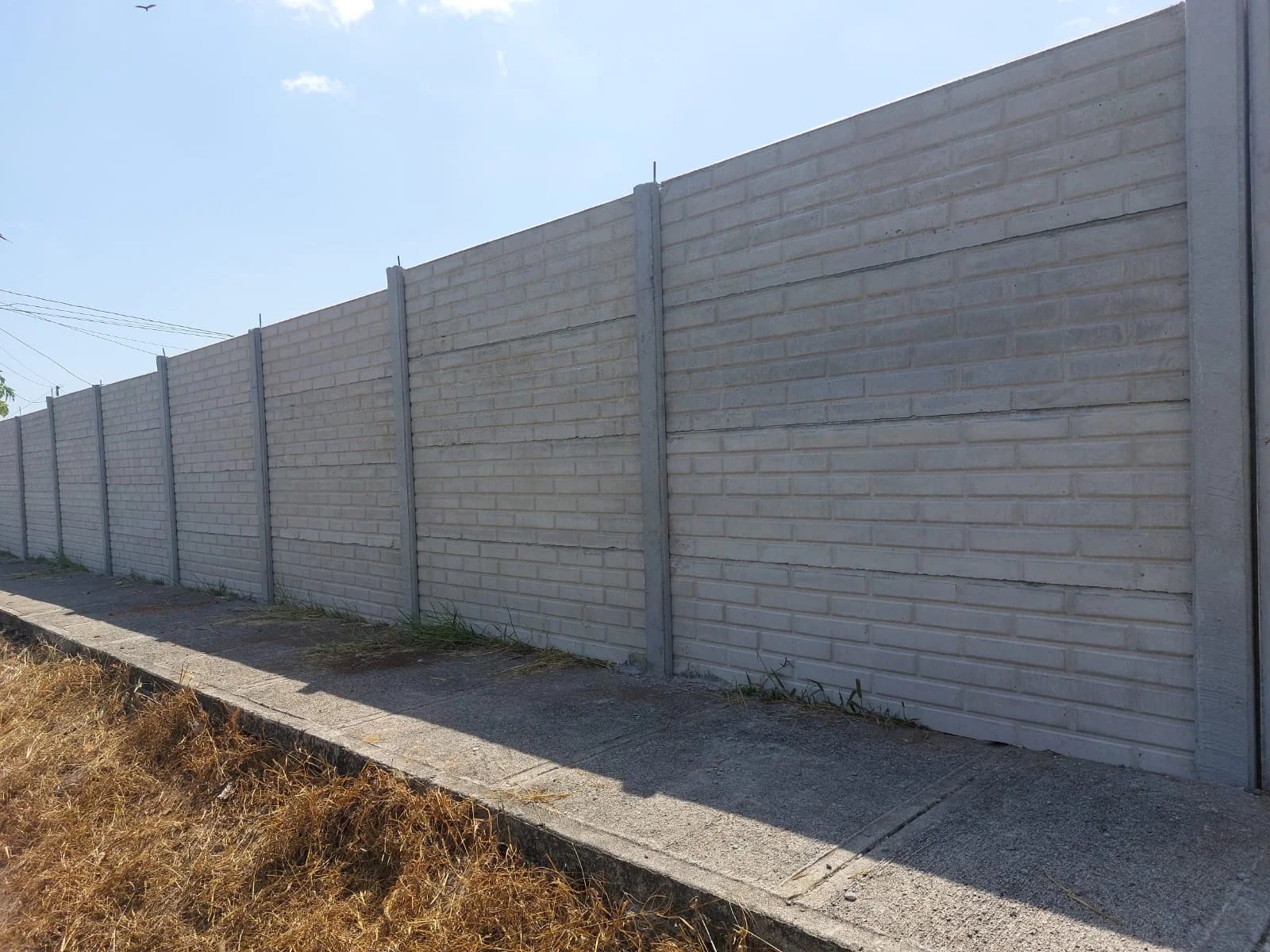 Muro perimetral prefabricado largo con textura tipo bloque y acera lateral en zona urbana