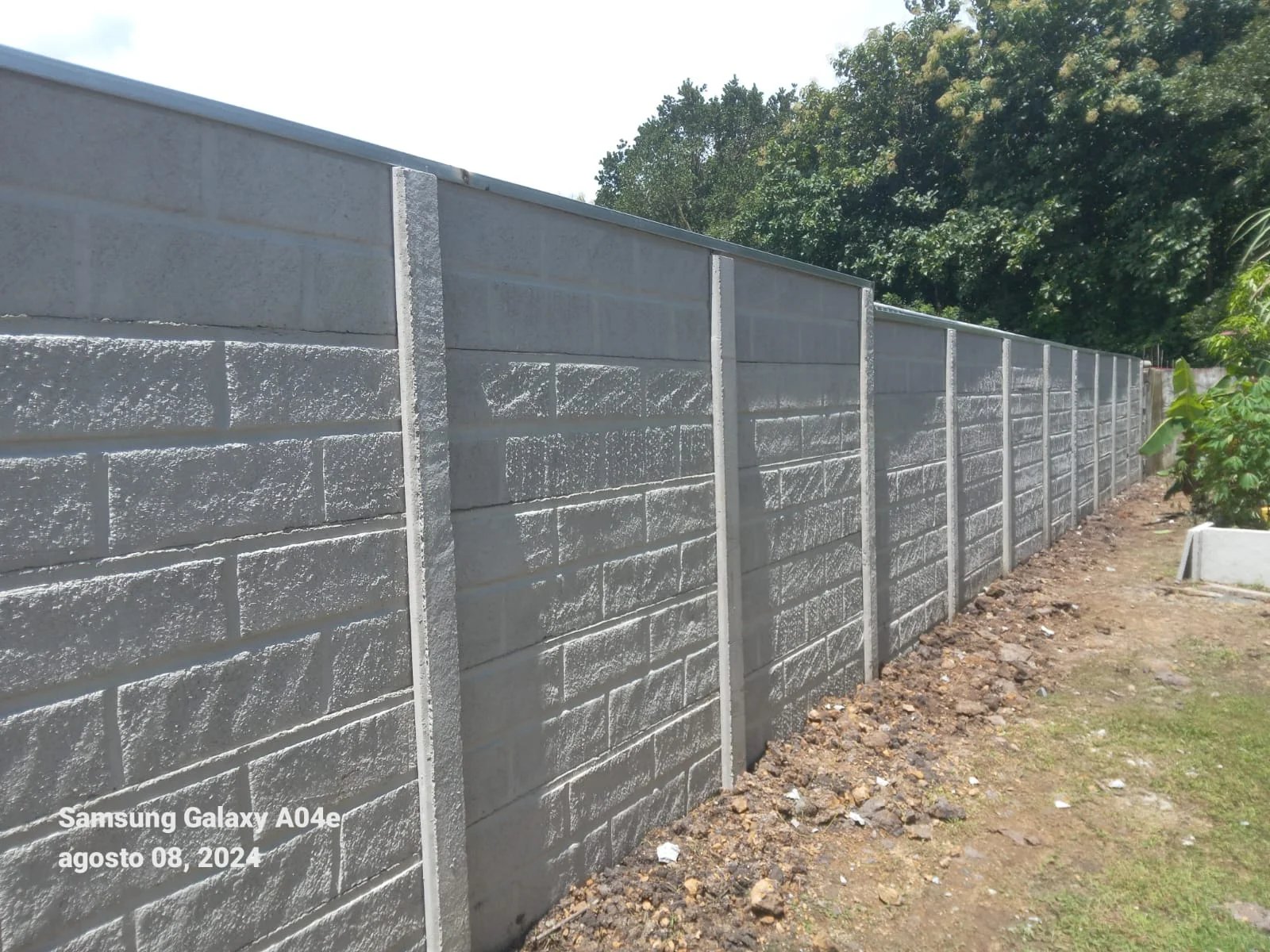 Muro perimetral prefabricado con textura tipo bloque gris en perspectiva larga junto a árboles