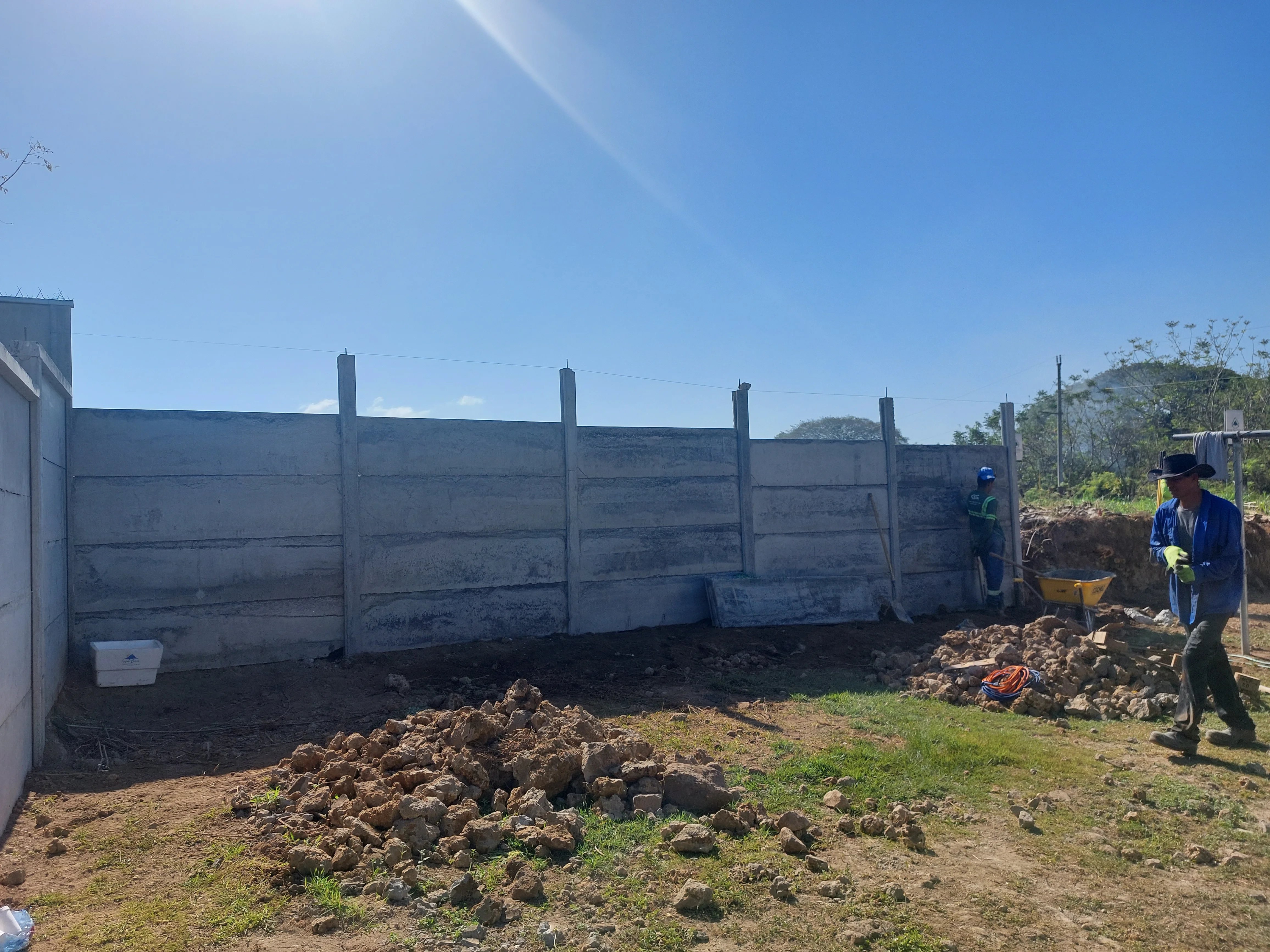 Muro perimetral prefabricado de concreto en lote rural con cuadrilla preparando el terreno al fondo