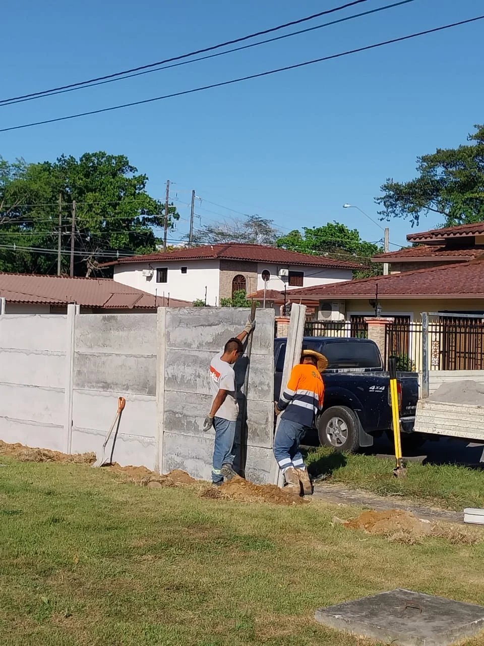Cuadrilla instalando muro perimetral en obra residencial con vivienda y vehículo al fondo